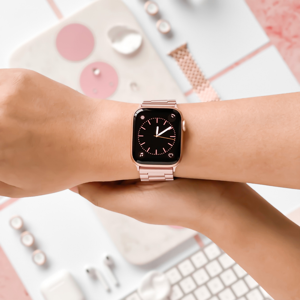 A Stainless Steel Apple Watch Strap - Blush by Coconut Lane is showcased on a person's wrist. The background features a matching aesthetic with a white keyboard, mouse, and various accessories in pastel shades, creating a clean, stylish setup.