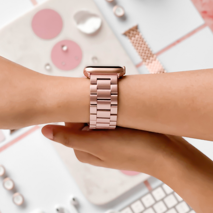 A close-up of a person's wrist wearing the Coconut Lane Stainless Steel Apple Watch Strap in Blush. The person is adjusting the strap with their other hand. The background features blurred desk items in pastel colors, including a keyboard, mouse, and stationery.